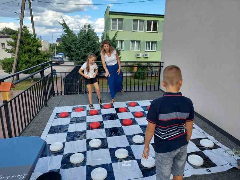 Taras biblioteczny, barierka, drzewa, ulica, trawnik, budynek banku, wielka plansza do gry, ludzie